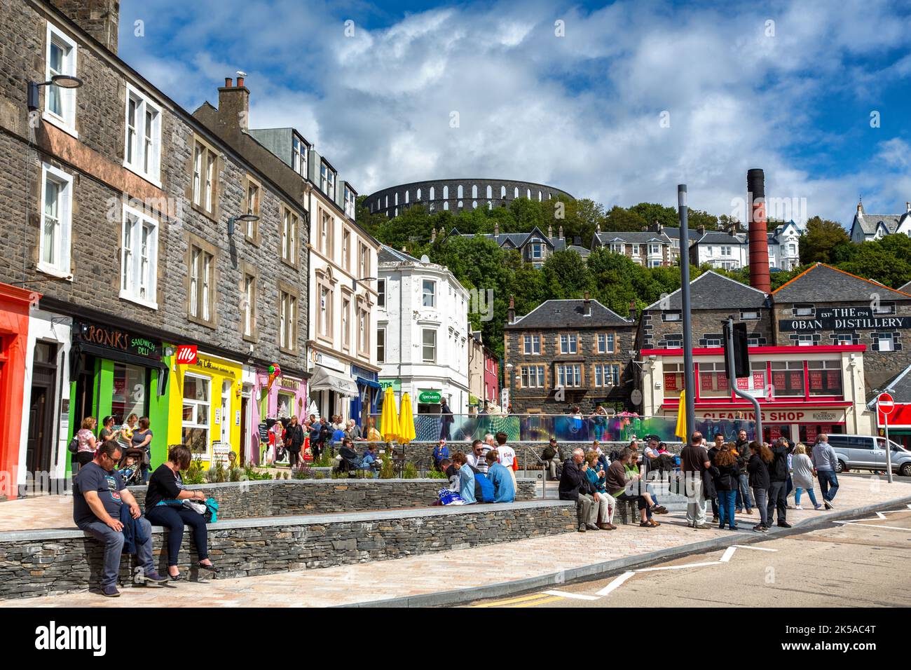 Oban, Scozia, Regno Unito - 29 luglio 2015: I colorati negozi e caffè sul lungomare di Oban, con i turisti che si godono una giornata estiva. Mostra i punti di riferimento locali di t Foto Stock