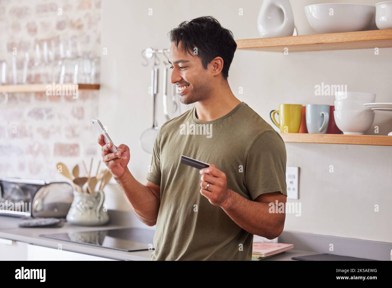 Carta di credito, finanza e uomo con smartphone per lo shopping online in cucina a casa. Indiano, faccia il pagamento con soldi per la transazione di e-commerce e. Foto Stock