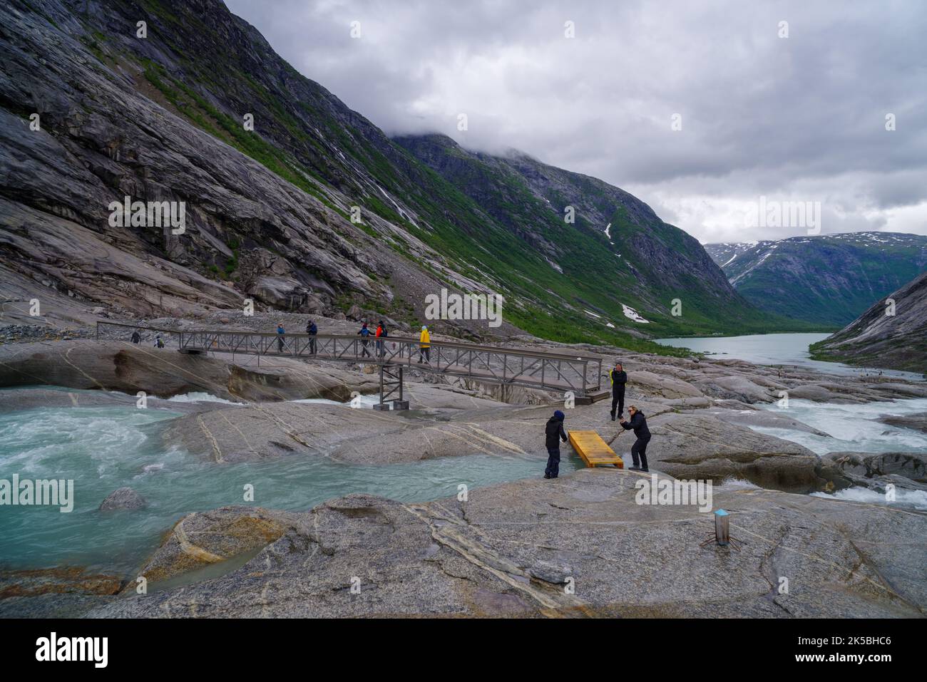 GJERDE, NORVEGIA - 3 LUGLIO 2022: Turisti che attraversano un ponte pedonale sulla loro strada per il ghiacciaio nigardsbreen sullo sfondo Foto Stock