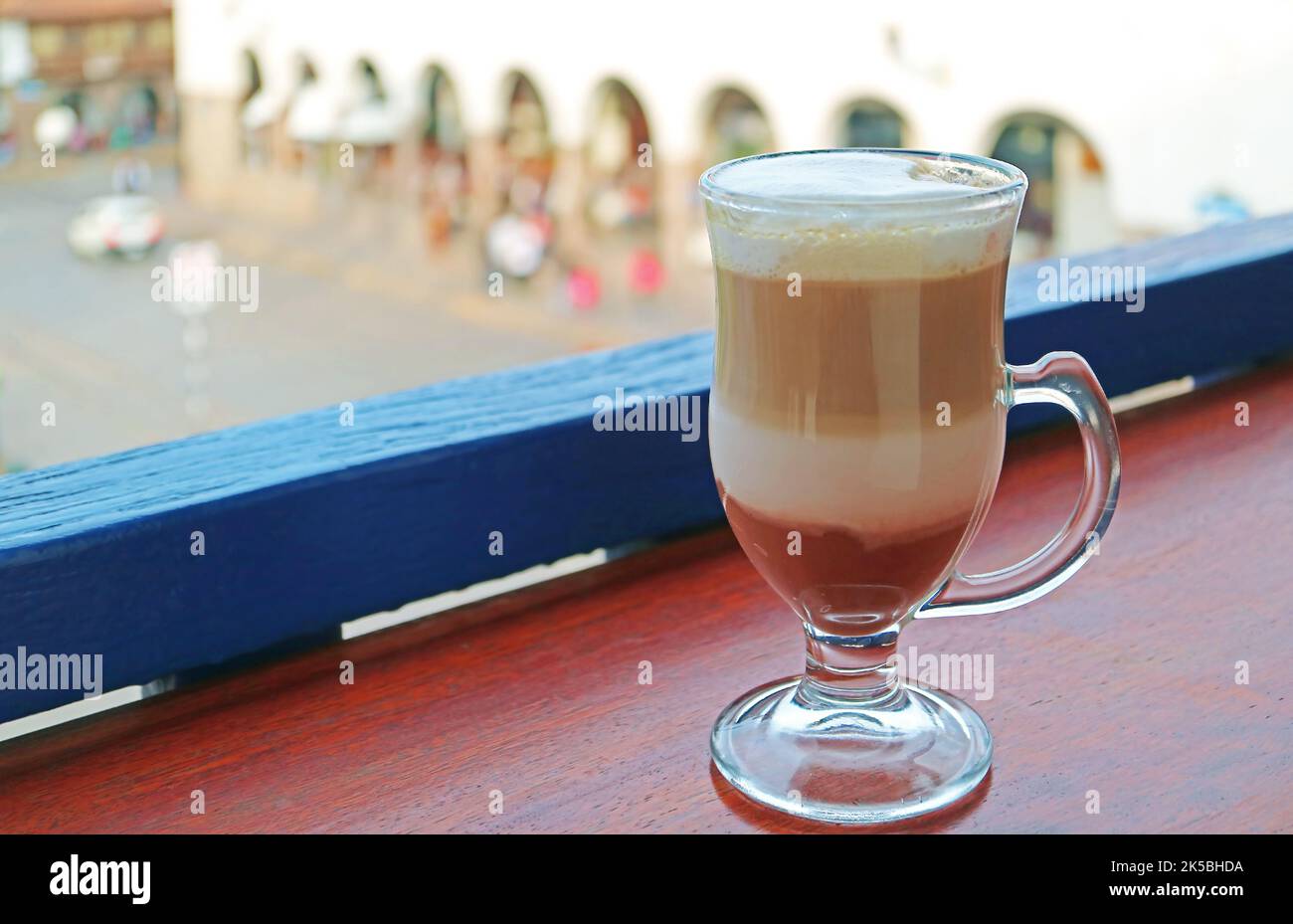 Una tazza di cappuccino caldo in una tazza trasparente con sfocata piazza della città vecchia di Cusco, Perù sullo sfondo Foto Stock