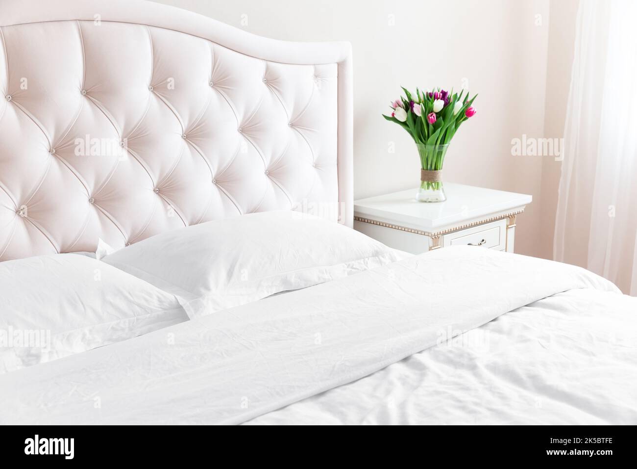 Camera da letto con letto, biancheria da letto bianca e comodino con bouquet di tulipani in vaso. Cuscini bianchi, piumone e piumone sul letto con testiera beige. Foto Stock