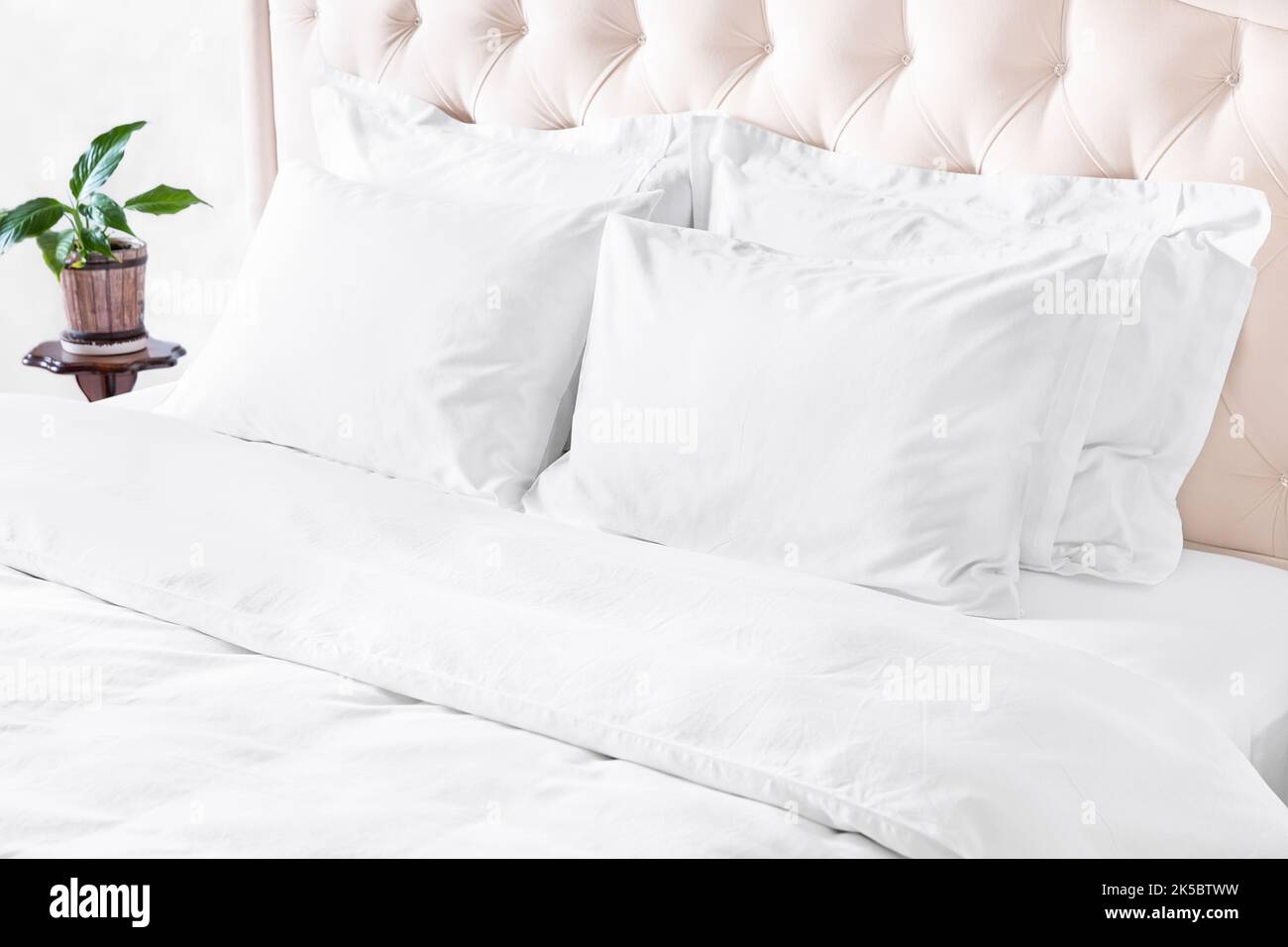 Camera da letto con letto, biancheria da letto bianca e comodino. Cuscini bianchi, piumone e copripiumino sul letto con testiera beige. Letto con cuscini bianchi puliti e. Foto Stock