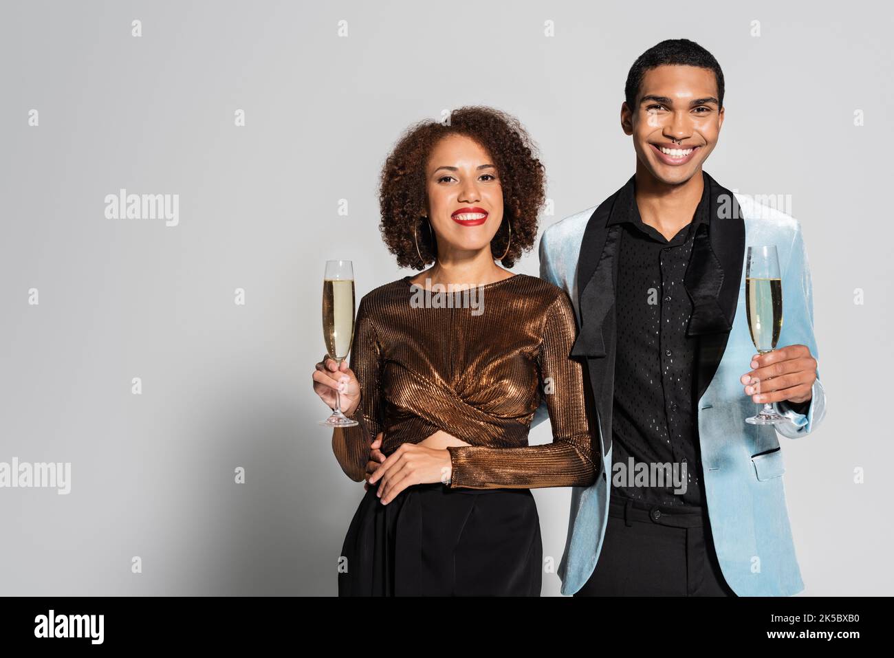 donna afroamericana in blusa lucida e uomo in blazer di seta con bicchieri da champagne su sfondo grigio, immagine stock Foto Stock