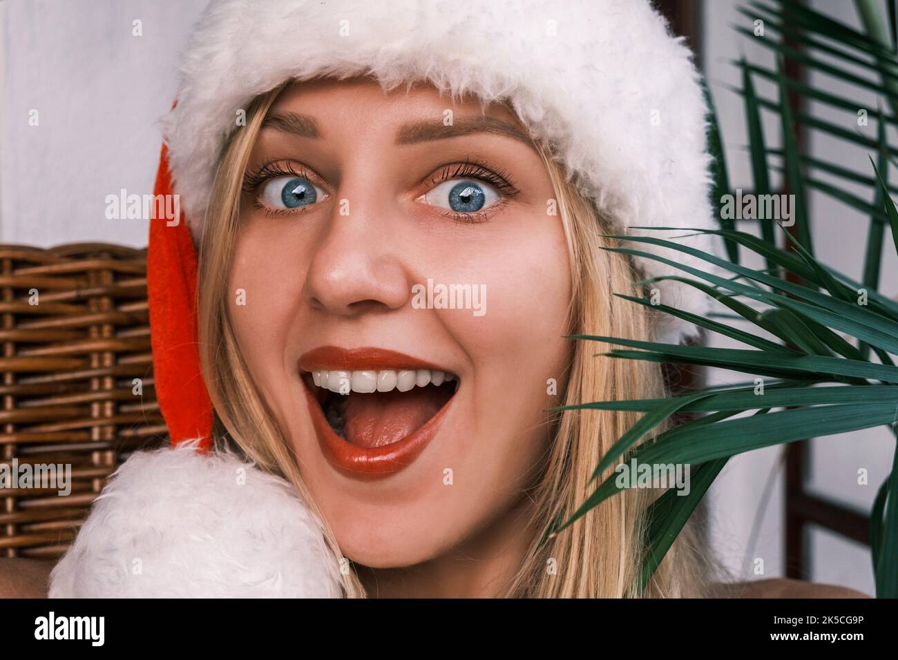 Divertente donna bionda sorridente che indossa il cappello di Santa guardando in macchina fotografica con gli occhi sorpresi. Ragazza che celebra il Natale nei tropici. Atmosfera festiva Foto Stock
