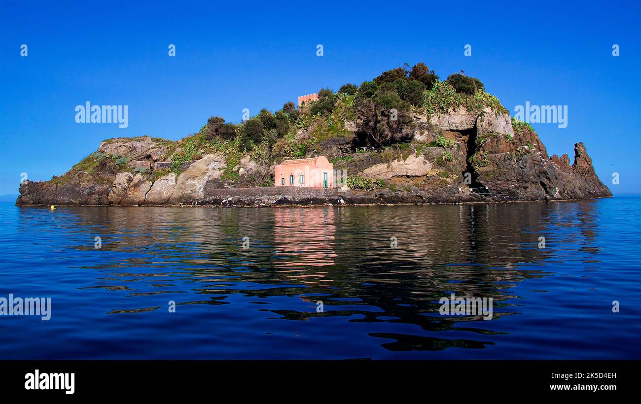 Italia, Sicilia, Costa dei Ciclopi, Isola dei Ciclopi, piccola isola boschiva, edificio rosa, mare scuro e calmo, cielo blu, senza nuvole Foto Stock