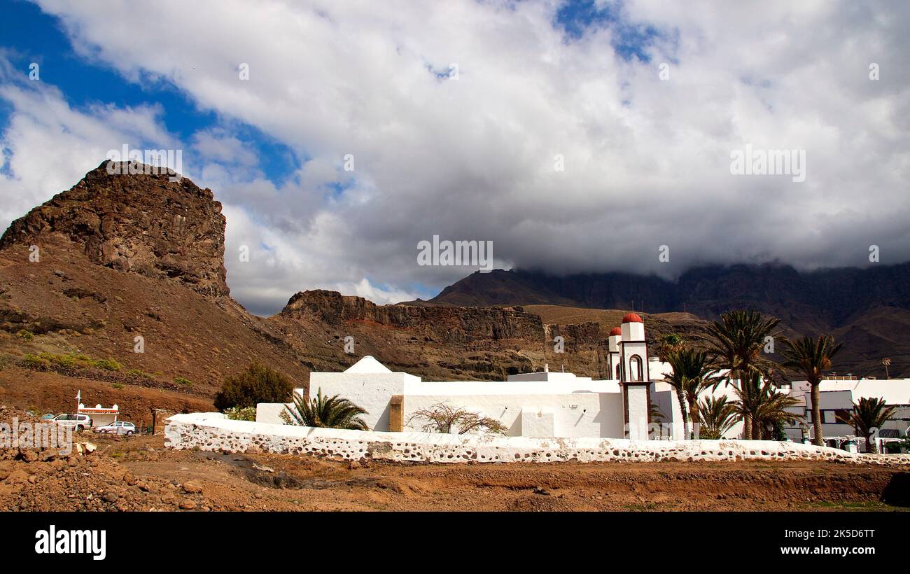 Spagna, Isole Canarie, Gran Canaria, costa occidentale, città portuale, Puerto de las Nieves, cielo blu, nuvole bianche di cumuli, eremo, Nuestra Senora de las Nieves, muro di pietra bianca, edificio bianco della chiesa, due campanili con tetti rossi, alte rocce sullo sfondo, palme sul terreno Foto Stock