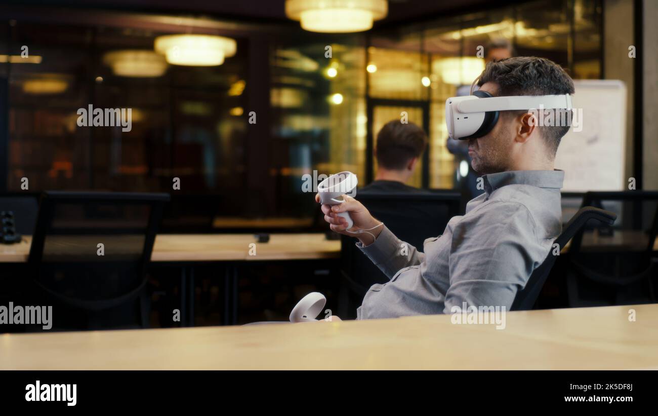 Vista posteriore di un uomo che indossa un visore VR e utilizza controller wireless, guardando i dati in realtà virtuale. Lavora in un ufficio moderno di un'azienda hi-tech con colleghi diversi. Tecnologia digitale nel cyberspazio Foto Stock