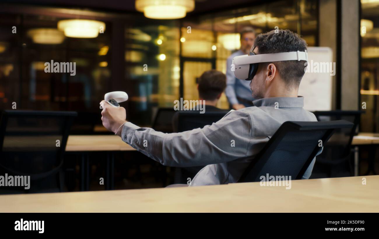 Vista posteriore di un uomo che indossa un visore VR e utilizza controller wireless, guardando i dati in realtà virtuale. Lavora in un ufficio moderno di un'azienda hi-tech con colleghi diversi. Tecnologia digitale nel cyberspazio Foto Stock