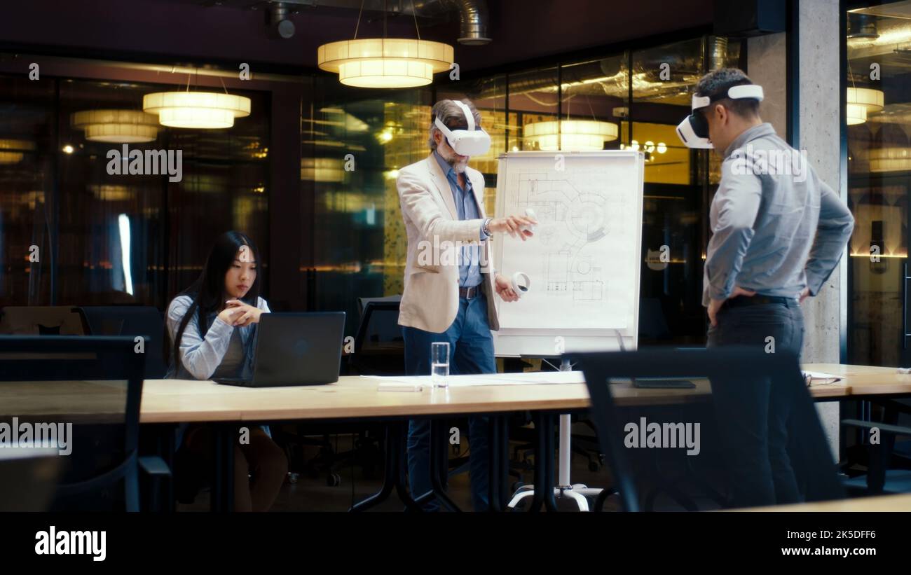 Ingegneri multietnici in visore VR, che utilizzano laptop e controller wireless, esaminano il progetto, guardano i dati, lavorano in uffici moderni di aziende hi-tech. Tecnologia digitale nel cyberspazio Foto Stock