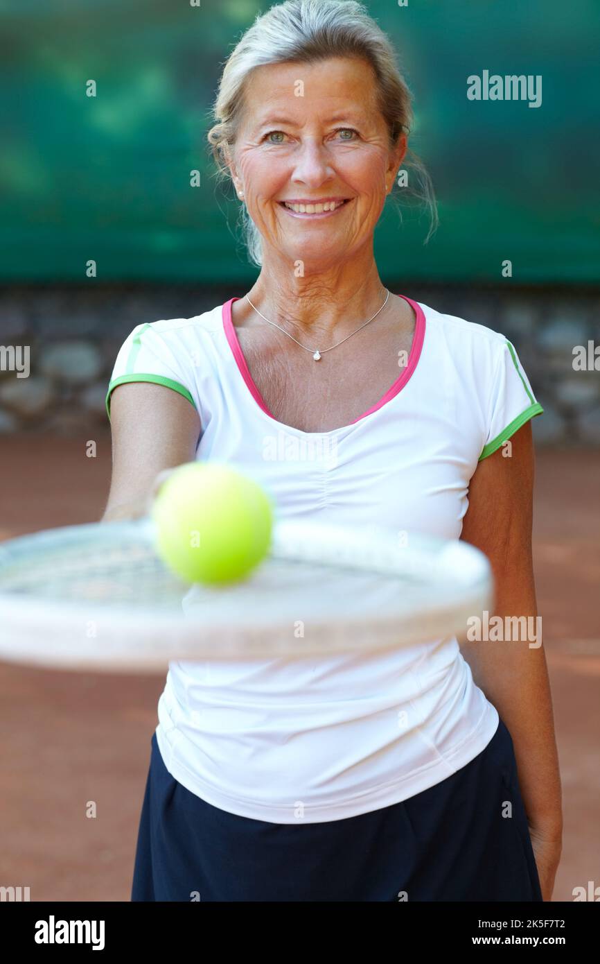 Lascia giocare un po' di tennis. Donna anziana sorridente mentre tiene fuori una racchetta da tennis con una sfera sulla parte superiore. Foto Stock