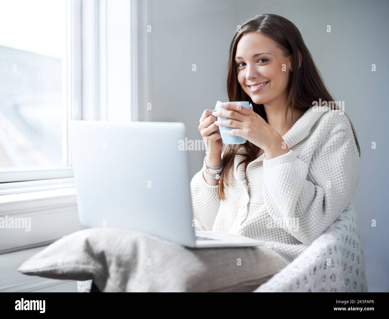 Intrattenimento al coperto per quei mesi invernali. Bella giovane brunetta sorseggiando un caffè mentre si siede a casa davanti al suo computer portatile. Foto Stock