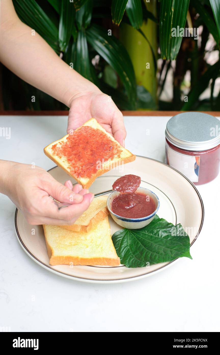 Mani spalmare la confettura di prugne su un pane tostato contro il vaso con la marmellata Foto Stock