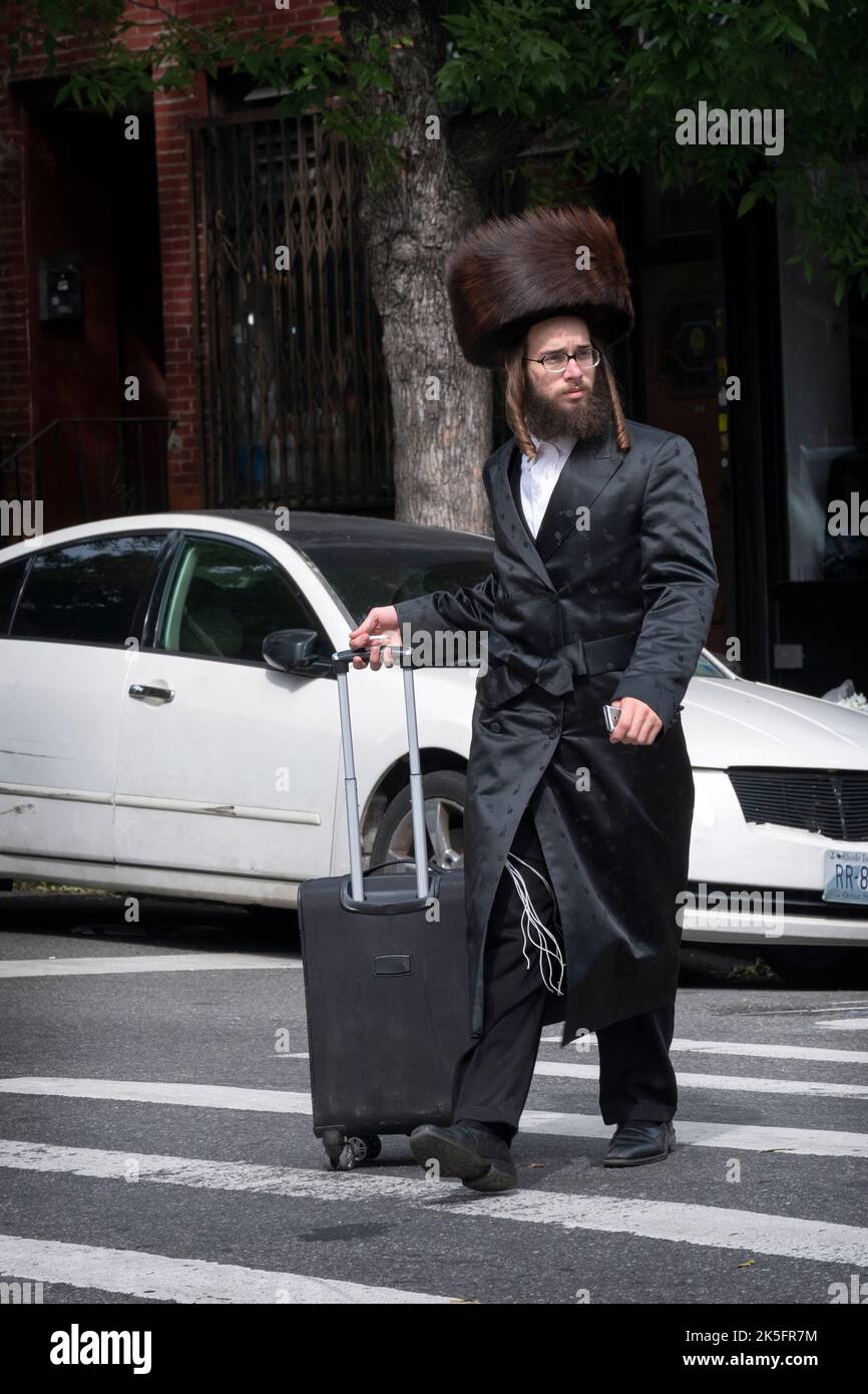 Un uomo ebraico ortodosso che si prepara al Sabbath indossa un cappello di pelliccia di shtreimel e attraversa la strada facendo una valigia. A Williamsburg, Brooklyn, New York. Foto Stock