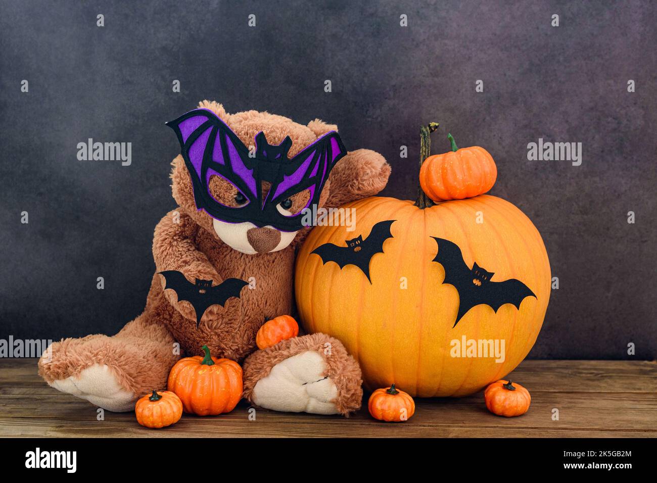 Divertente orsacchiotto marrone con maschera a forma di pipistrello e zucche su vecchio tavolo di legno da parete nera Foto Stock