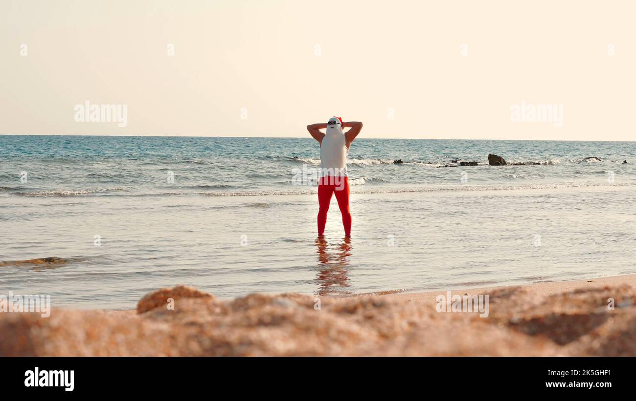 Babbo Natale si bagna il sole. Babbo Natale divertendosi. Babbo Natale divertente, in occhiali da sole, in piedi in acqua di mare sulla spiaggia. Vacanze estive a Babbo Natale. Foto di alta qualità Foto Stock