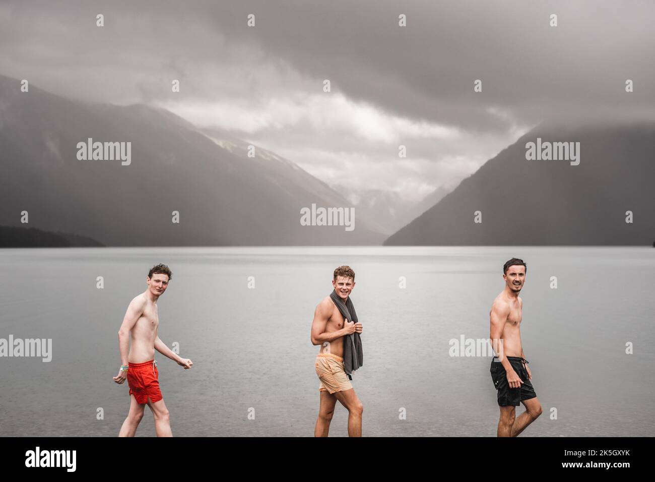 tre giovani uomini caucasici che guardano la macchina fotografica mentre camminano da sinistra a destra lungo la costa fredda e calma del lago d'acqua in tronchi di nuoto in mezzo alla natura in solitaria Foto Stock