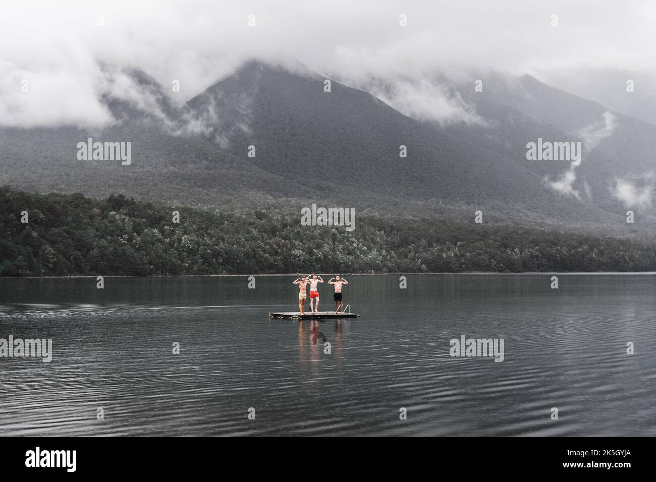 tre ragazzi caucasici in piedi vestiti da nuoto che scherzano dalla piattaforma di legno nel centro del lago nelle acque calme nel mezzo di Foto Stock