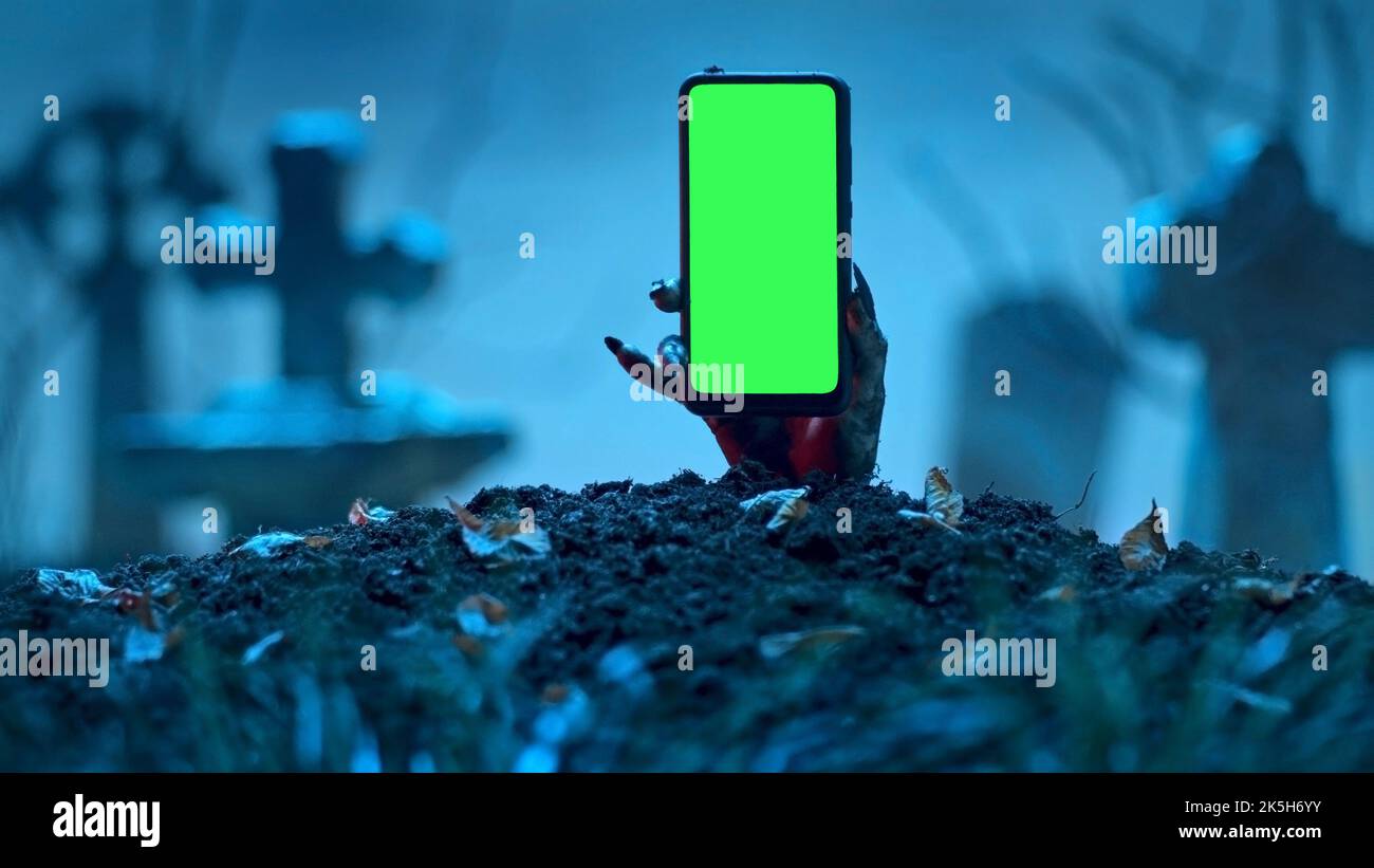 Mano zombie che si alza smartphone con schermo verde fuori dalla tomba. Evento di festa Halloween concetto. Foto Stock