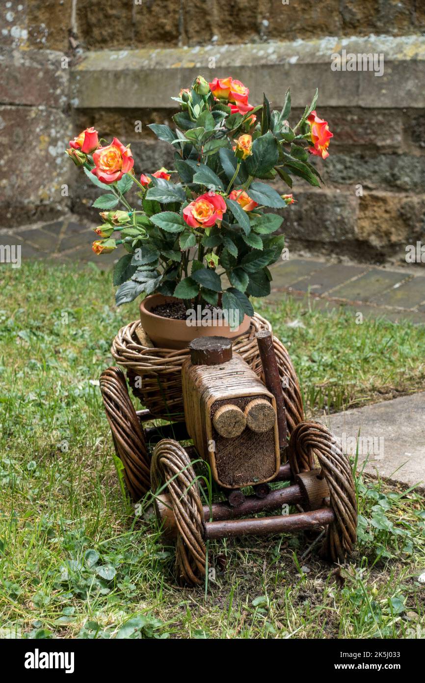 Trattore ornato in vimini con piantatrice a lato del cimitero, piccolo cespuglio di rose e fiori rossi, Little Dalby Church, Leicestershire, Inghilterra, Regno Unito Foto Stock