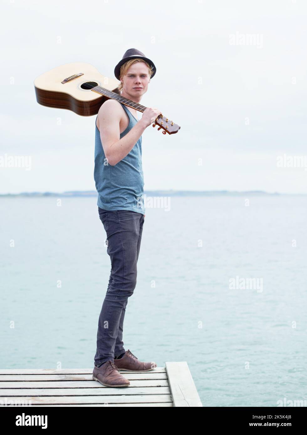 Sempre con la chitarra. Un'immagine a tutta lunghezza di un giovane elegante che porta una chitarra e si trova in piedi su un molo vicino all'acqua. Foto Stock