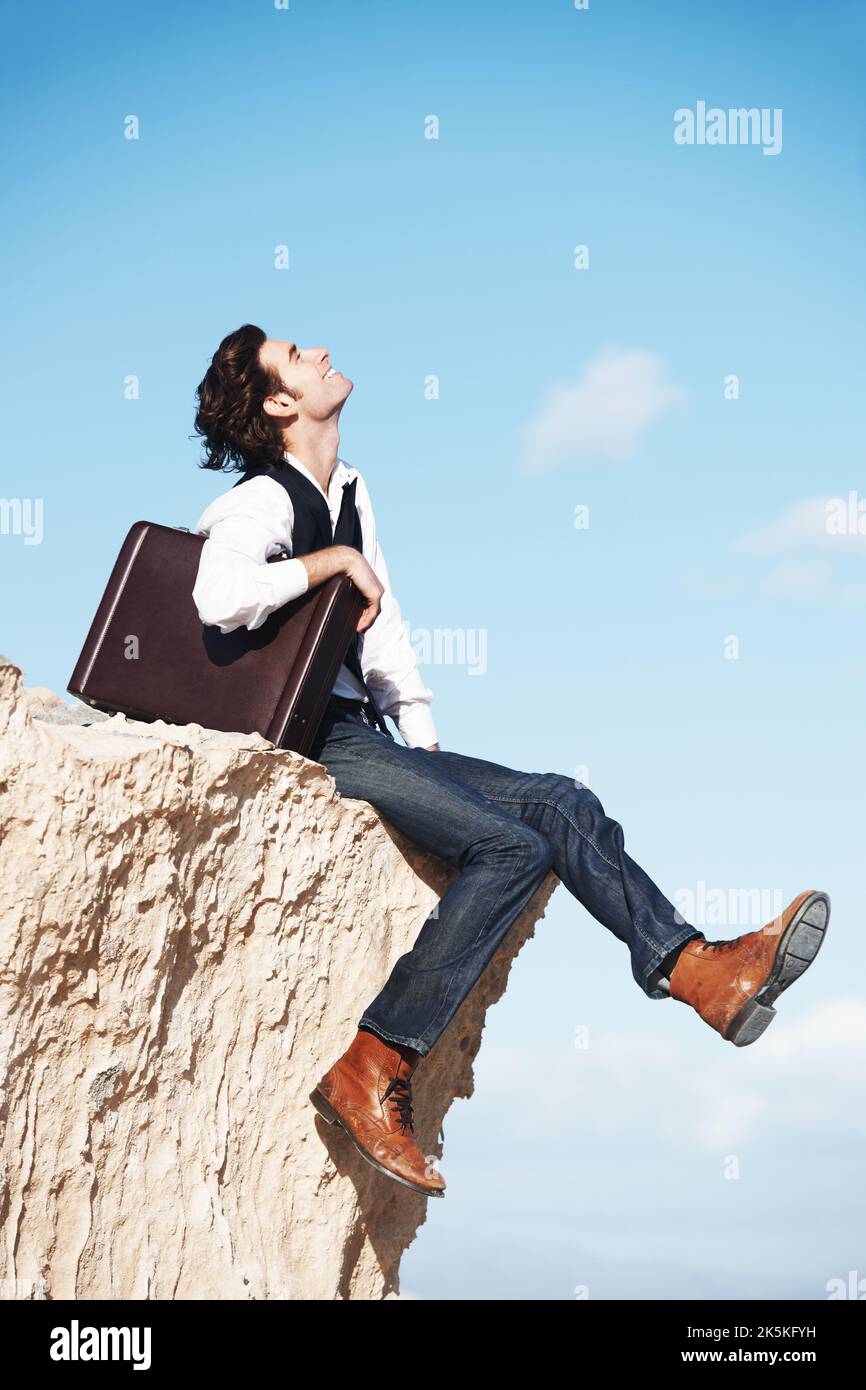 Godendo di un po' di aria fresca. Giovane uomo sorridente seduto con la sua valigetta sul bordo di una scogliera che domina l'oceano. Foto Stock