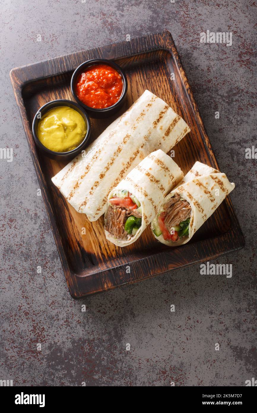 Doner kebab a base di carne cotta su una rotisserie verticale con aggiunte di verdure e salsa avvolta in closeup lavash sul bordo di legno sulla t Foto Stock