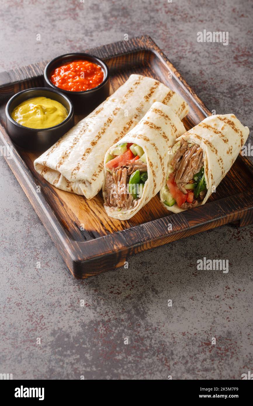 Doner kebab a base di carne cotta su una rotisserie verticale con aggiunte di verdure e salsa avvolta in closeup lavash sul bordo di legno sulla t Foto Stock