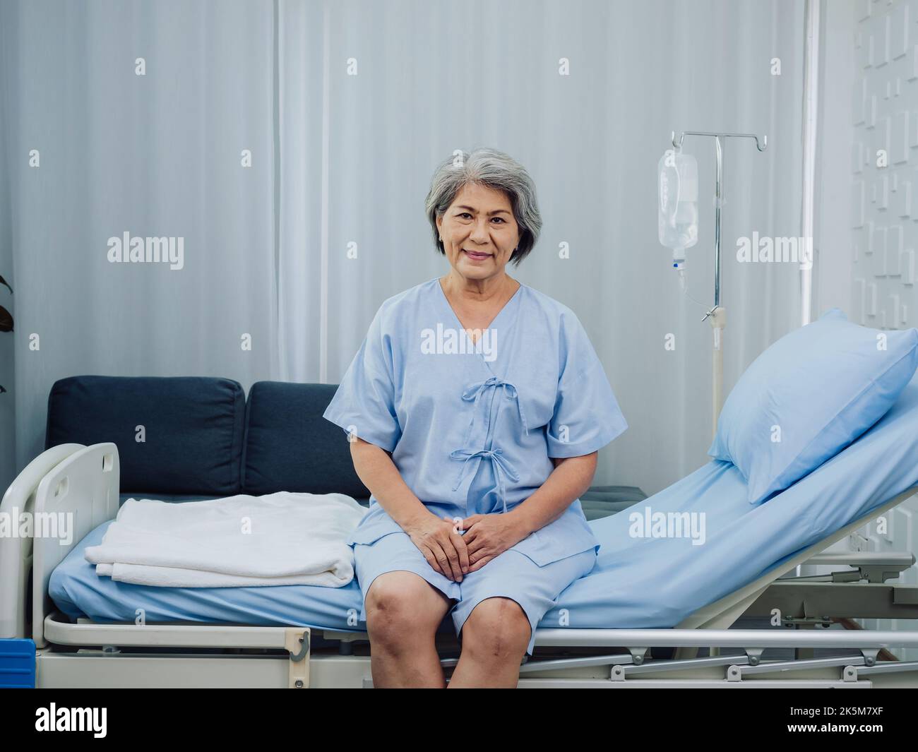 Felice sorriso bella asiatica anziana paziente con chirurgia del ginocchio in vestito azzurro seduto sul letto vicino al palo medico IV stand con soluzione salina Foto Stock