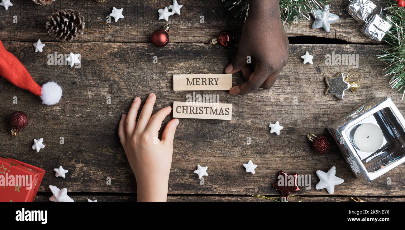 Mani di bambini caucasici e neri che fanno un buon segno di natale scritto su pioli di legno che li mette su scrivania rustica di legno piena di ornamento di festa Foto Stock