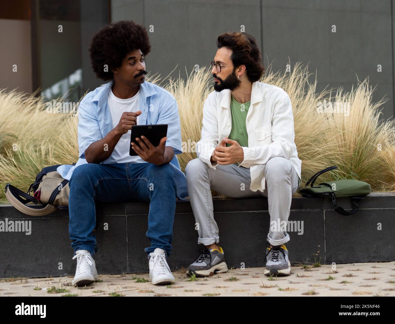 Due giovani uomini diversi seduti in un quartiere finanziario mentre parlano e lavorano su un tablet. Lavoro di squadra Foto Stock
