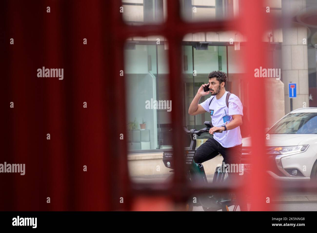 LONDRA - 21 maggio 2022: L'uomo usa il telefono cellulare mentre noleggiasse una bicicletta elettrica. Vista tramite la tradizionale cassetta telefonica Foto Stock