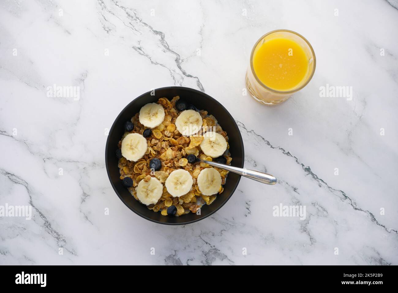 Colazione sana di grani interi con latte, mirtilli, banane e succo d'arancia su banco di marmo bianco. Foto Stock