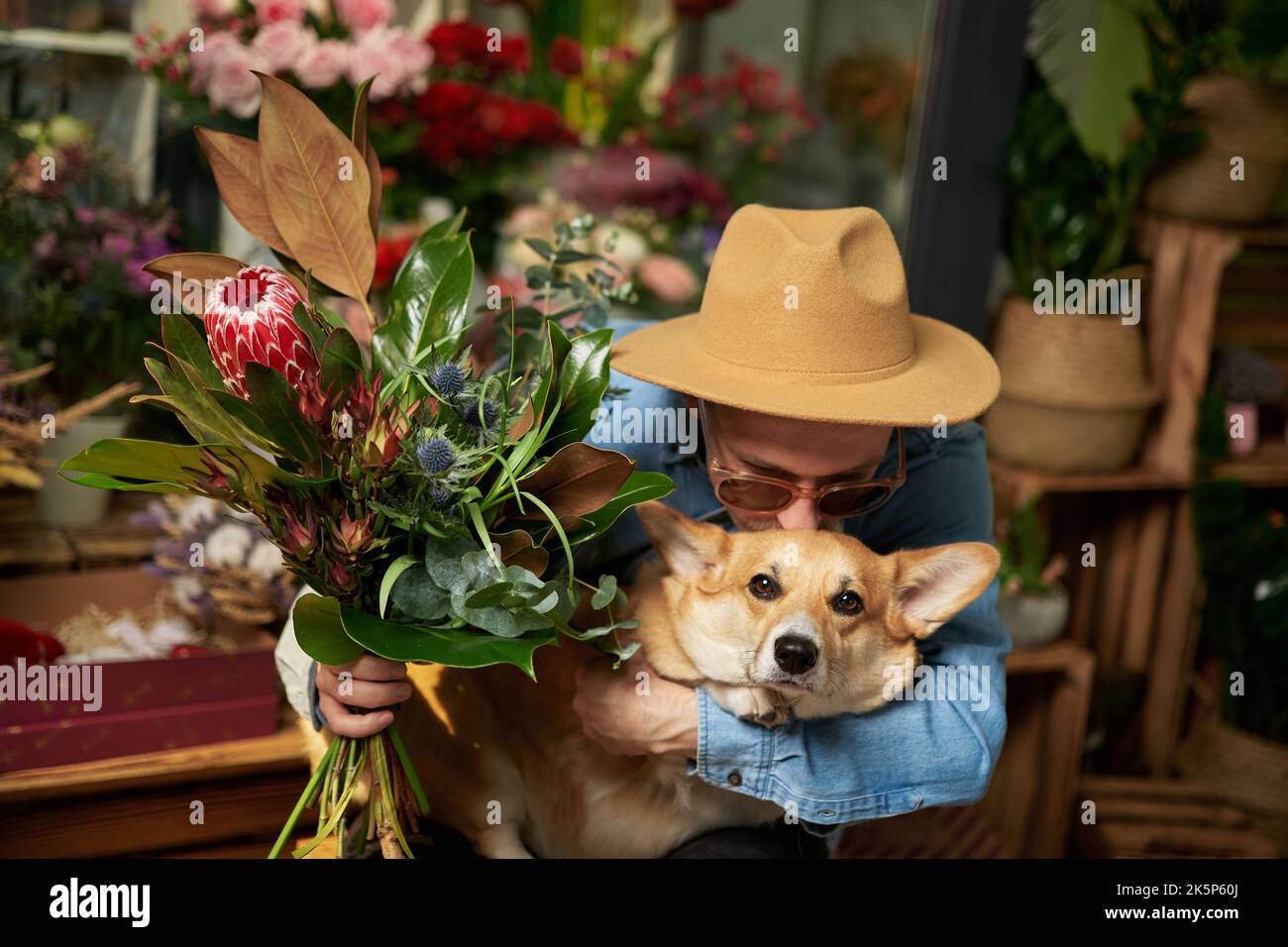 Hippster uomo in occhiali da sole e cappello con bouquet di fiori freschi di primavera baciare il cane corgi gallese. Giorno della mamma, giorno di San Valentino o Giornata internazionale della donna. Immagini di alta qualità Foto Stock
