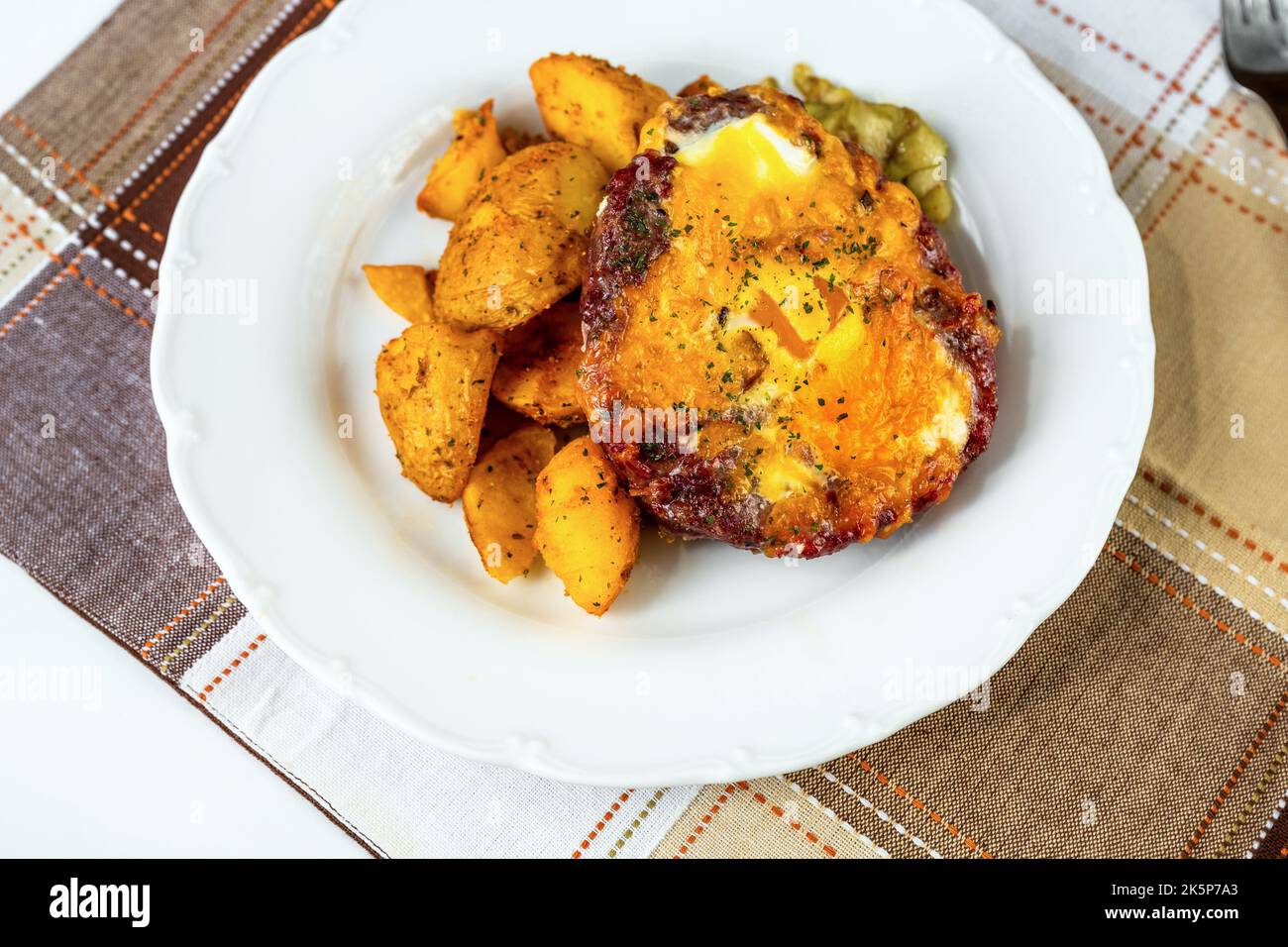 Hamburger di manzo con uova fritte, patate al forno e cubetter sottaceto su piatto bianco su tavolo a scacchi, primo piano. Foto Stock
