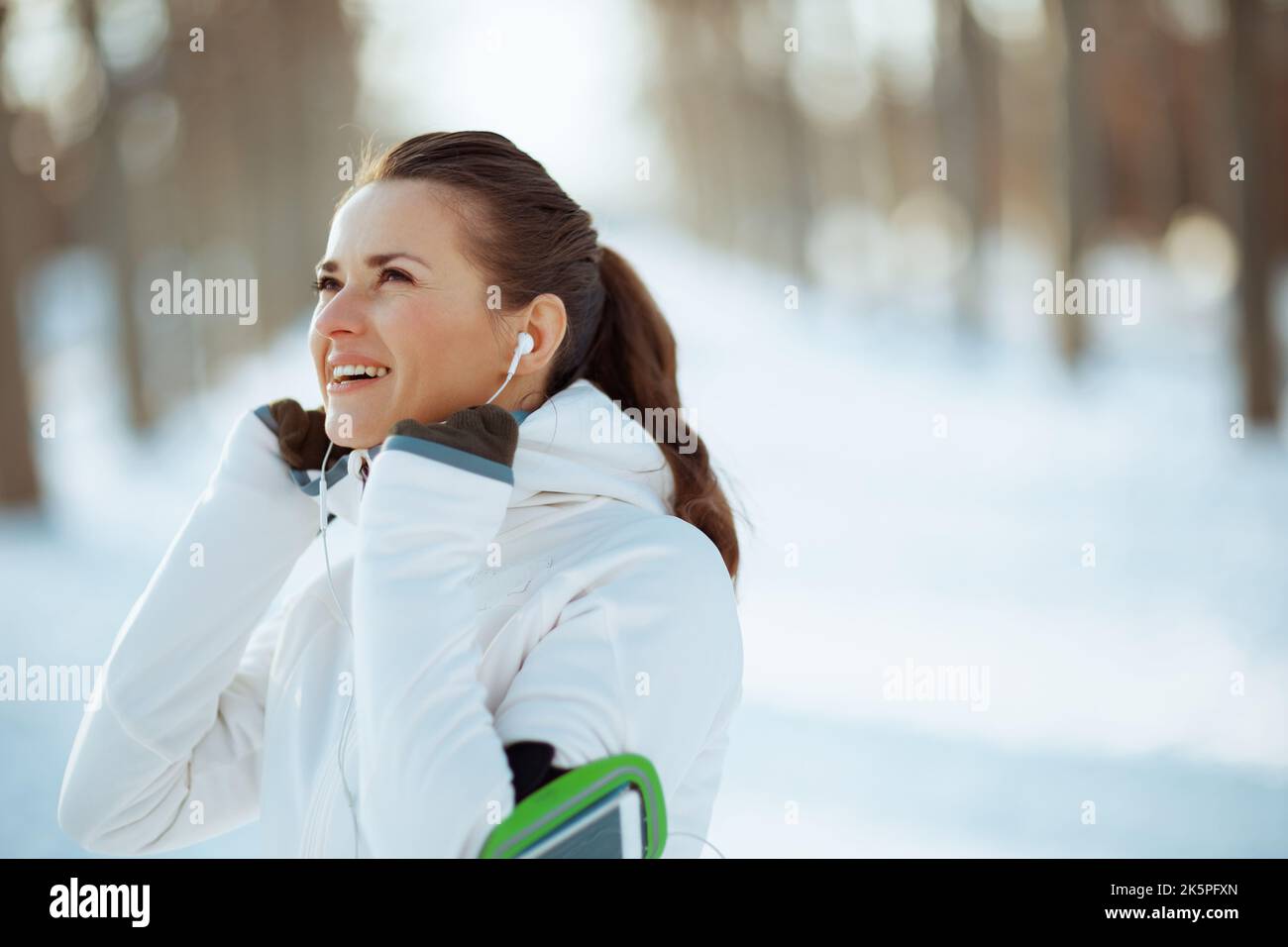 donna fitness sorridente in giacca bianca con cuffie all'aperto nel parco cittadino in inverno. Foto Stock