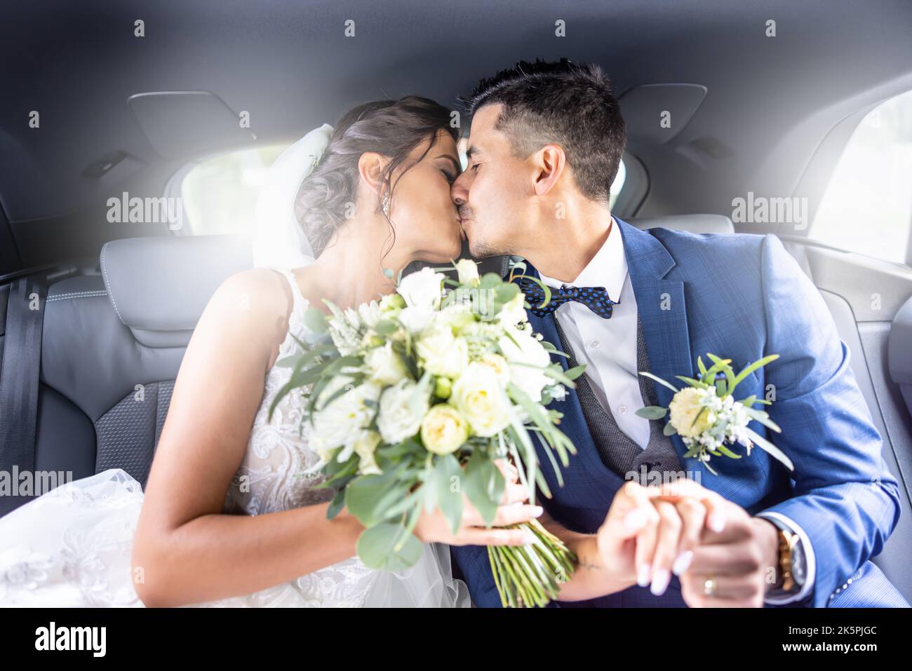 Felice giovane appena sposato coppia bacia in macchina tenendo le mani e sposa in possesso di un bouquet di nozze. Foto Stock