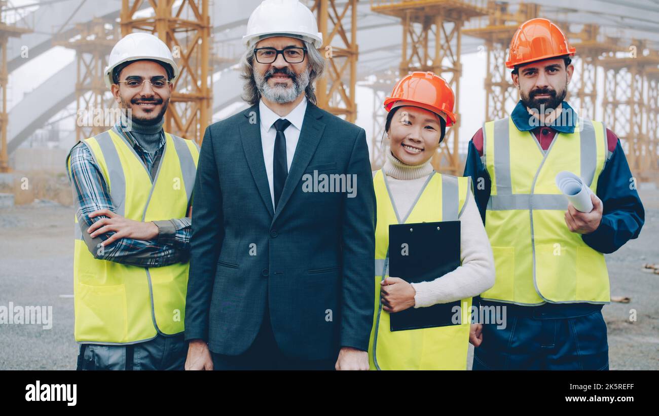 Il team multirazziale di architetti di successo sta in piedi all'aperto nel sito del progetto sorridendo tenendo documenti e progetti. Persone e concetto di occupazione. Foto Stock
