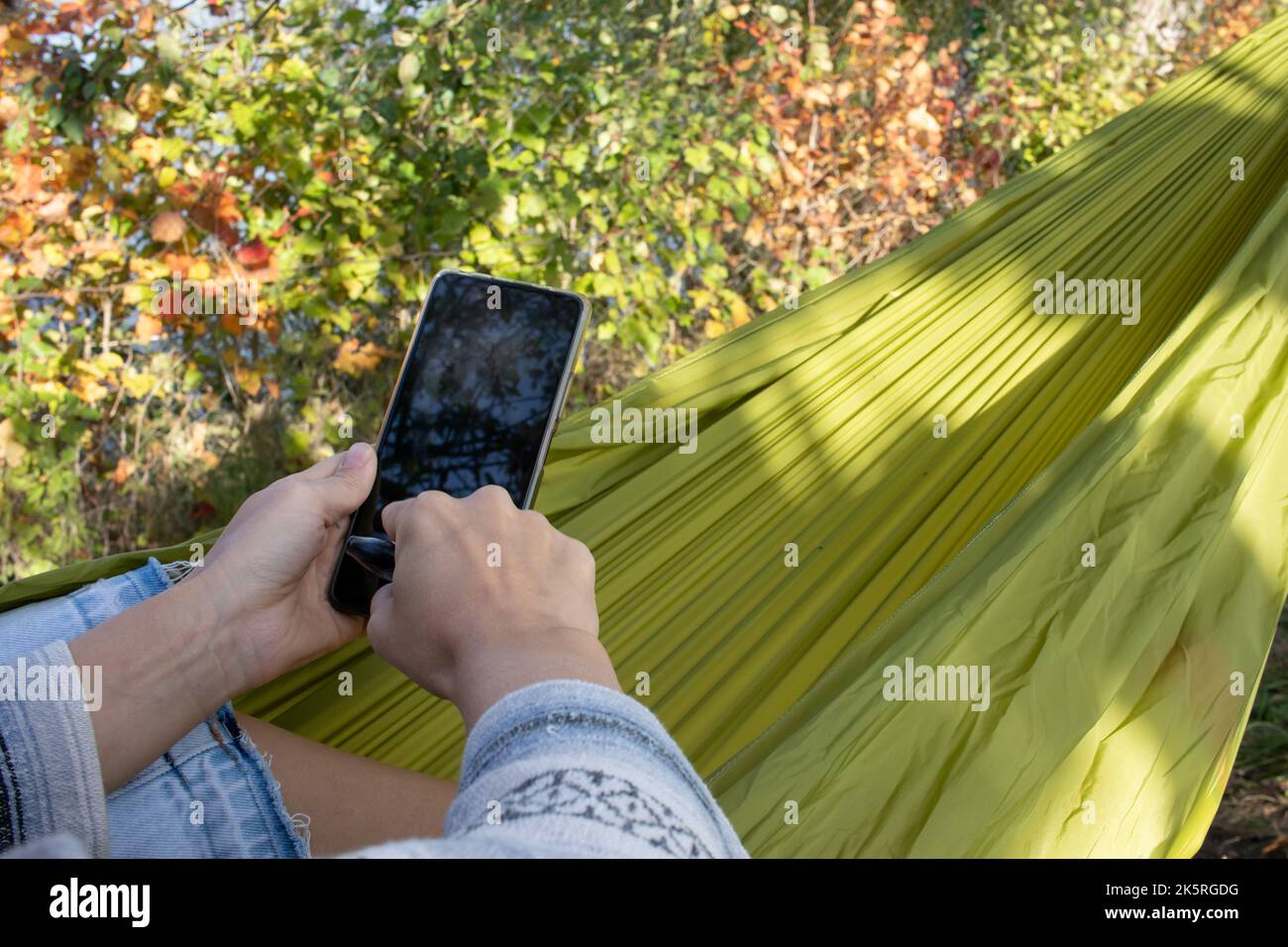 Giovane donna che si rilassa in amaca all'aperto in natura e con uno smartphone. Le mani tengono in mano uno smartphone e digitano un messaggio Foto Stock