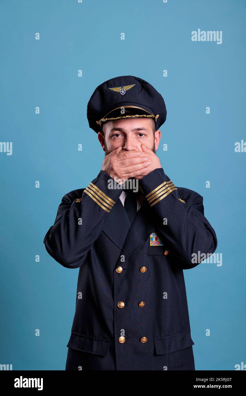 Pilota di aeroplano che copre la bocca con le mani, mostrando parlare non male gesto, guardando la fotocamera. Capitano aereo dell'accademia dell'aviazione con vista frontale delle labbra coperte, censura, mantenimento del concetto di silenzio Foto Stock