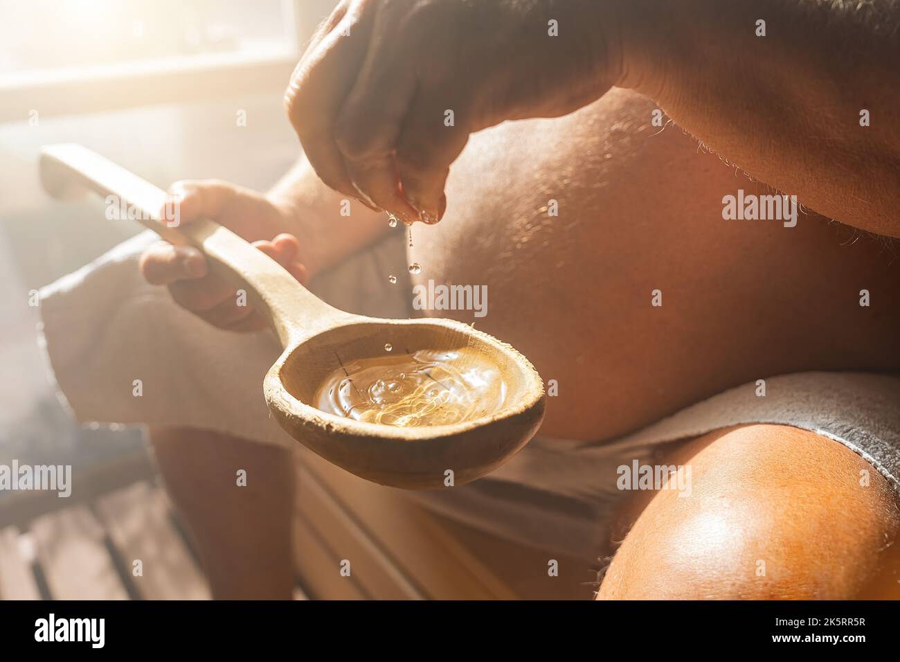 L'uomo anziano che gocciola acqua in un vecchio cucchiaio di legno nella sauna finlandese, spa e temperatura calda bagno terapia immagine concetto. Foto Stock