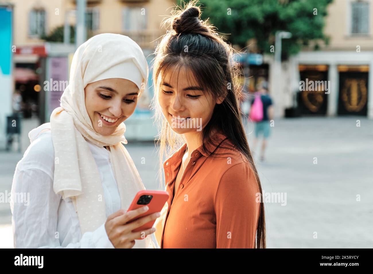Amici di sesso femminile multietnico sorridenti mentre usano un telefono cellulare insieme in piedi all'aperto per strada. Foto Stock