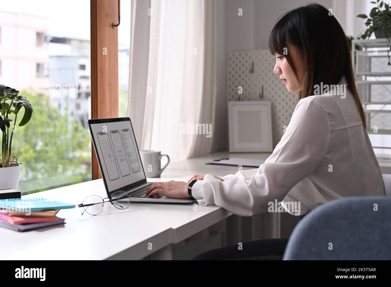 Vista laterale della giovane donna asiatica sviluppatore web che lavora con prototipo wireframe per web telefono cellulare sullo schermo del computer Foto Stock