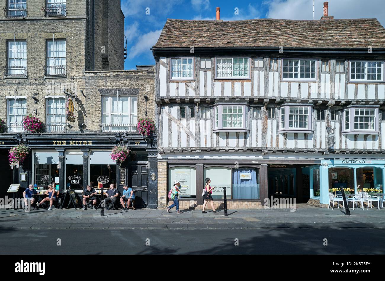 I locali si siedono fuori da un pub bevendo alcolici in una calda giornata estiva, mentre i turisti passano davanti a un negozio di abbigliamento, Bridge Street, Cambridge, Inghilterra. Foto Stock