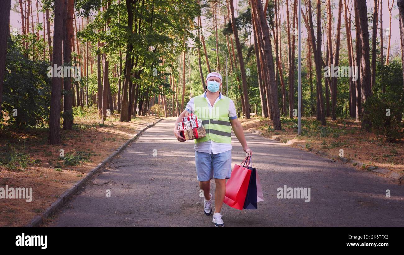 uomo, corriere, in maschera protettiva, tenendo in mano diverse confezioni di regali e borse per la spesa. all'aperto. consegna di regali per eventi di festa. donazione, concetto di beneficenza. Foto di alta qualità Foto Stock