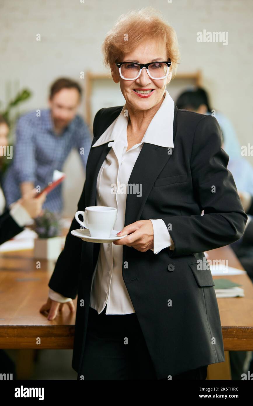 Ritratto di donna d'affari anziana e di successo. Donna felice e intelligente in tuta d'affari in un ufficio moderno durante un incontro con i colleghi Foto Stock