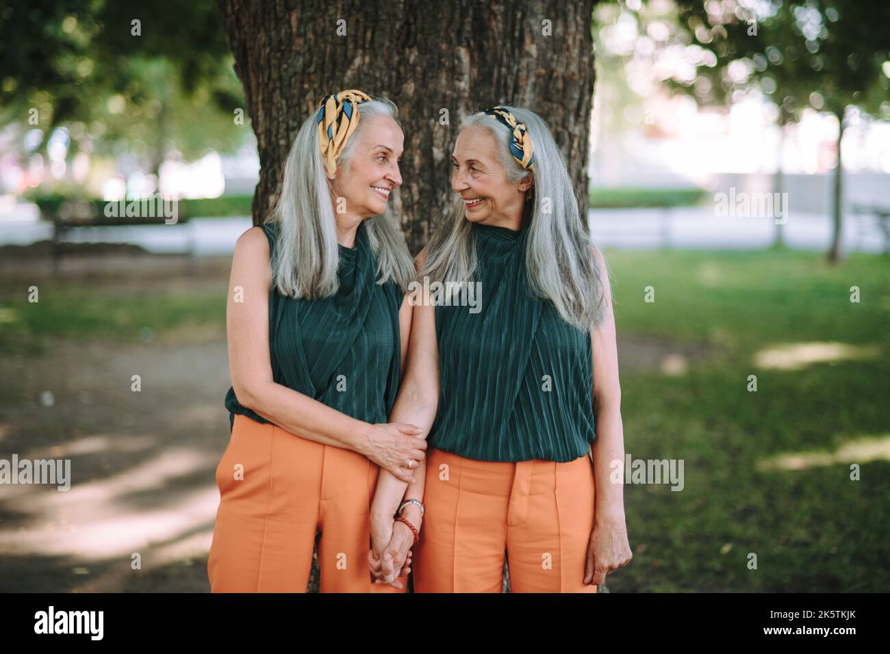 Donne anziane felici, gemelle che si posano davanti all'albero nel parco cittadino, sorridendo e parlando. Foto Stock