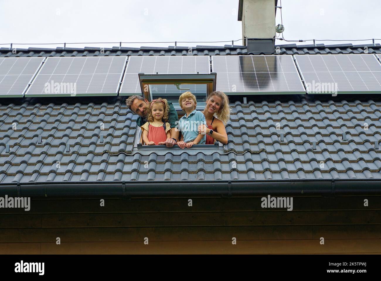 Famiglia felice che si appoggia fuori dalla finestra del lucernario nella loro nuova casa con pannelli solari sul tetto. Energia alternativa, risparmio di risorse e sostenibilità Foto Stock