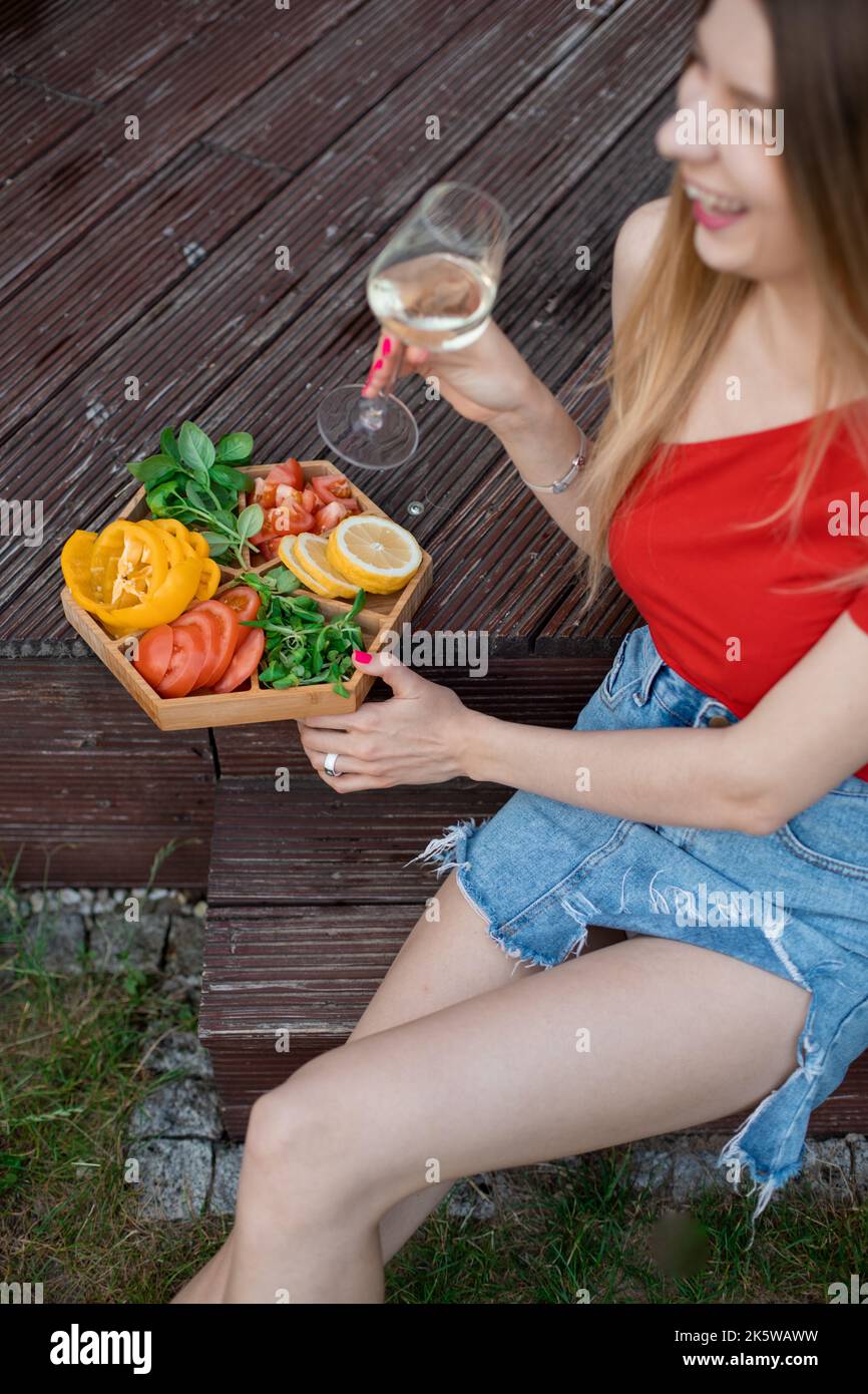 Donna bionda ridente verticale, studente che beve alcol e che mangia gustosi spuntini sani durante la festa. Vacanza in campagna Foto Stock