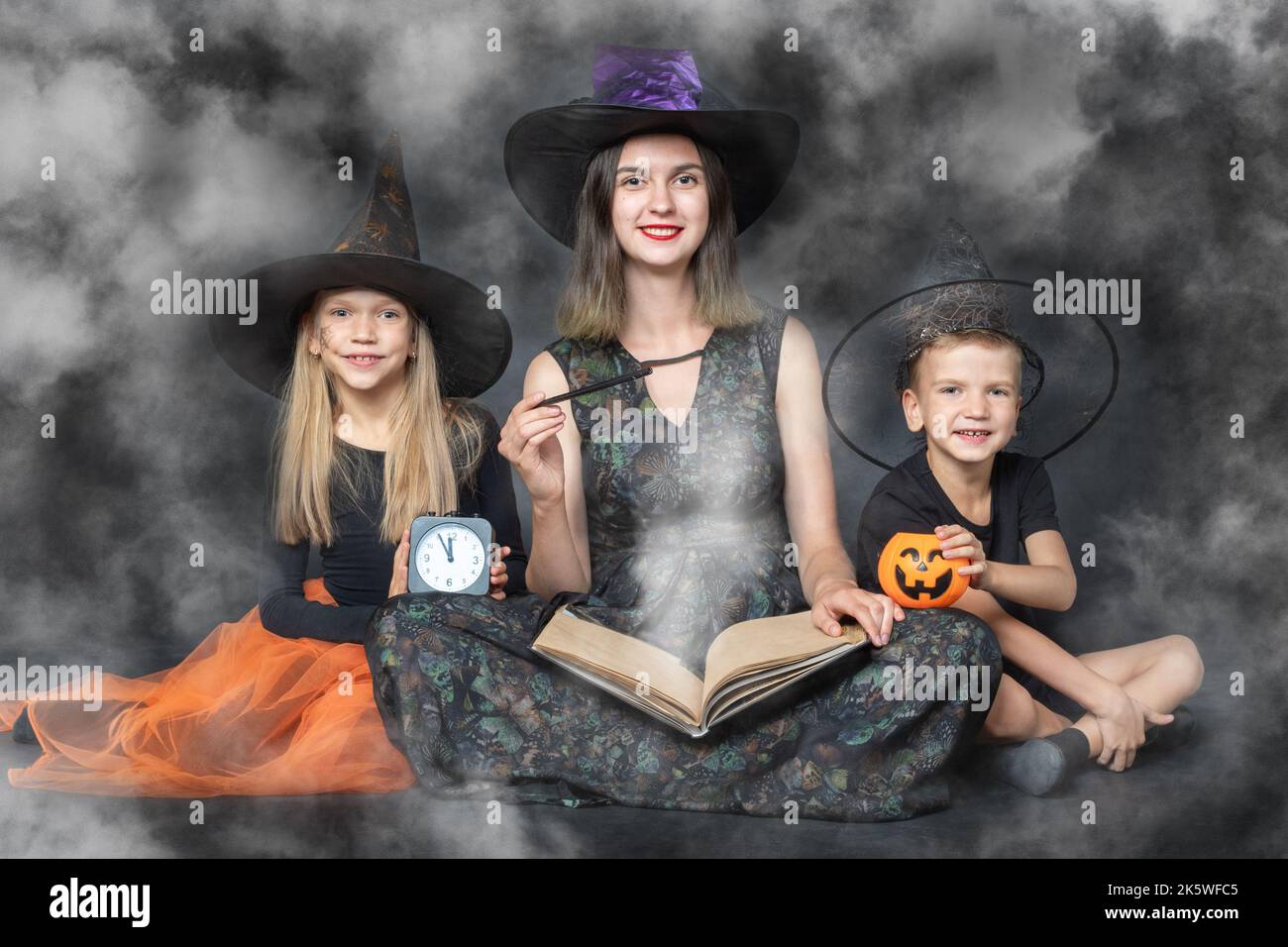 Una famiglia felice in costumi raccapriccianti di streghe e maghi, tenendo un grande libro con previsioni, cestini a forma di zucca, un orologio nero e un Foto Stock