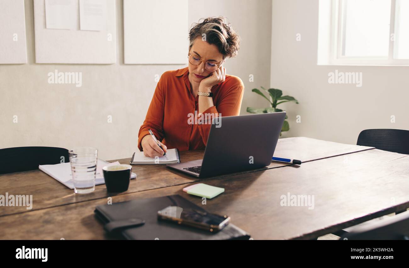 Donna d'affari matura che scrive nel suo diario mentre è seduta davanti a un laptop nel suo ufficio. Designer femminile scrivere la sua lista di cose da fare durante il lavoro Foto Stock